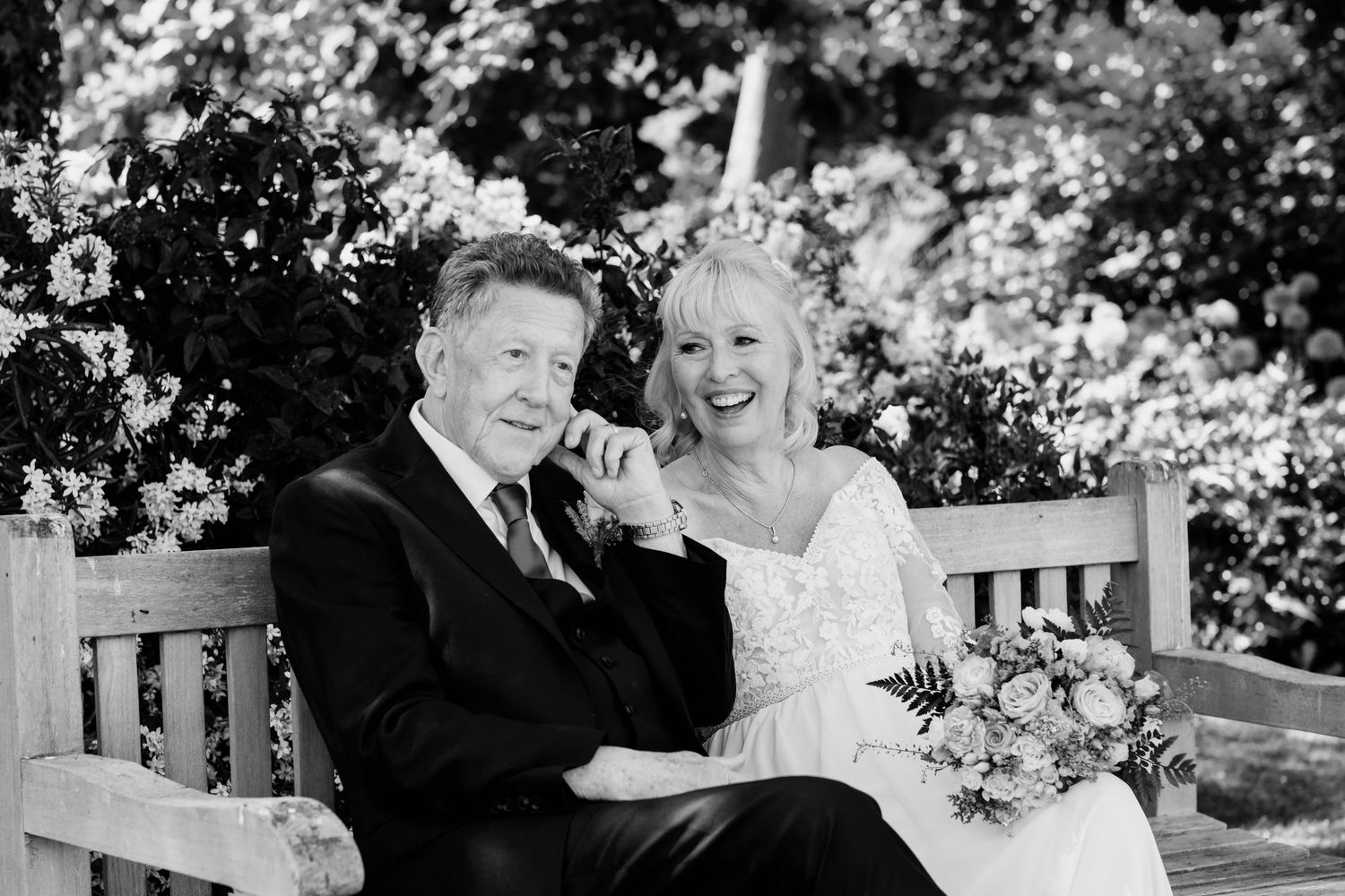 A black and white wedding photo of a couple sitting together on a wooden bench in a garden setting with flowers.