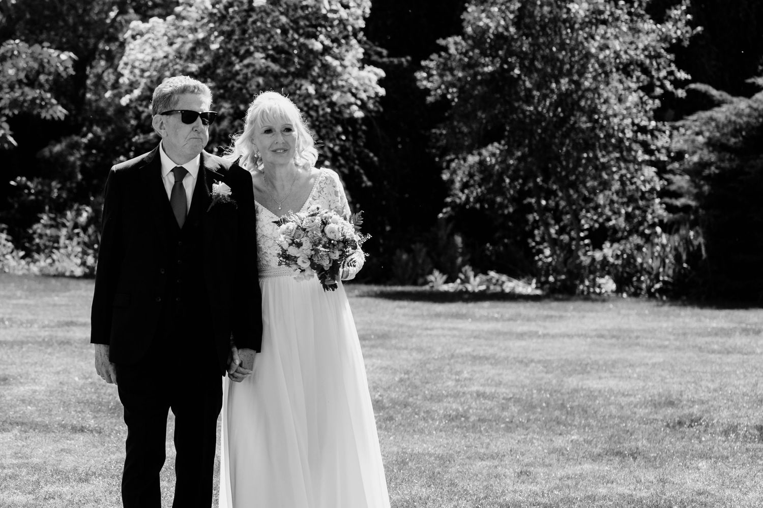 A black and white outdoor wedding photo of a couple standing together on a sunny lawn with trees in the background.