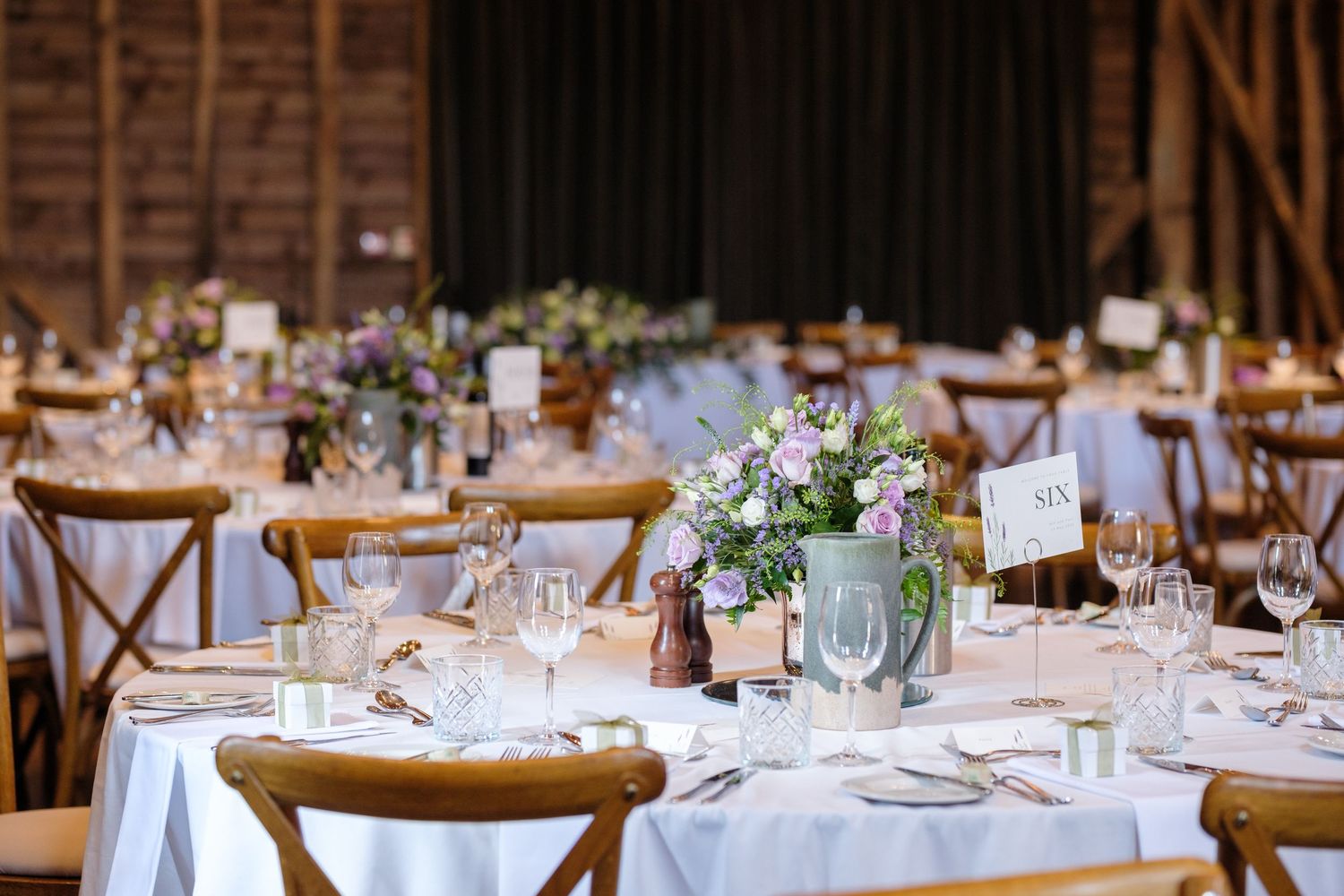Elegant rustic barn wedding reception with white tablecloths, wooden crossback chairs and delicate floral centerpieces.