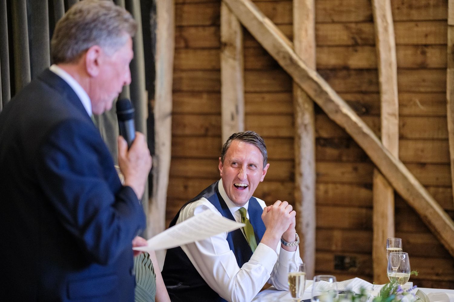 Two people share a candid laughing moment during speeches at a rustic wooden barn venue.