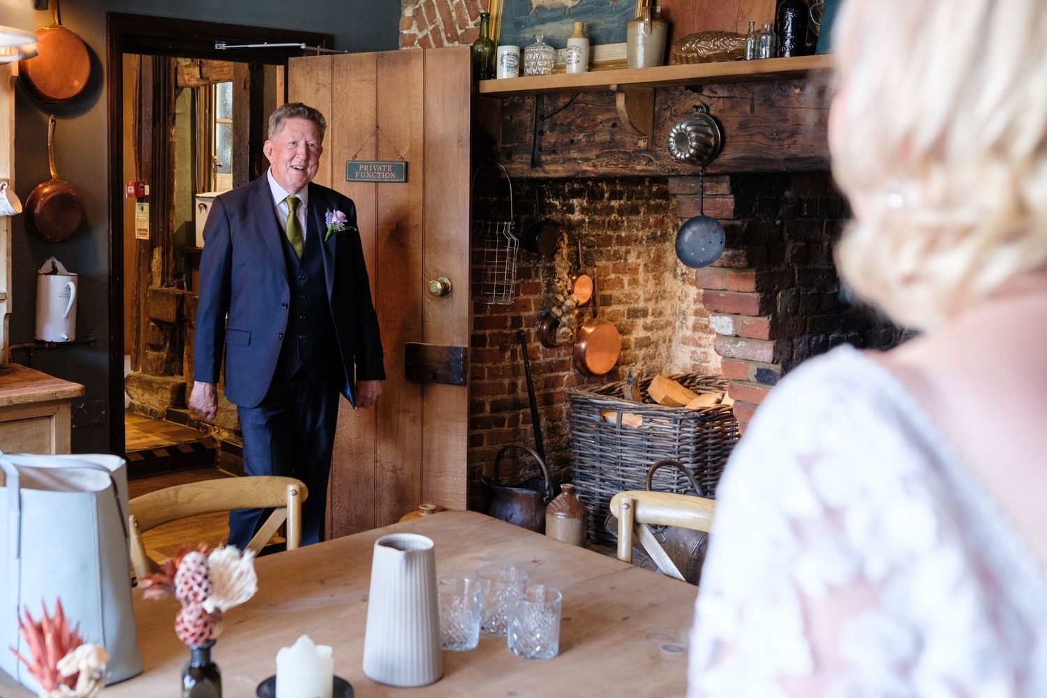 An elegantly dressed person in a blue suit is shown inside a rustic room with a vintage brick fireplace and kitchen decor.