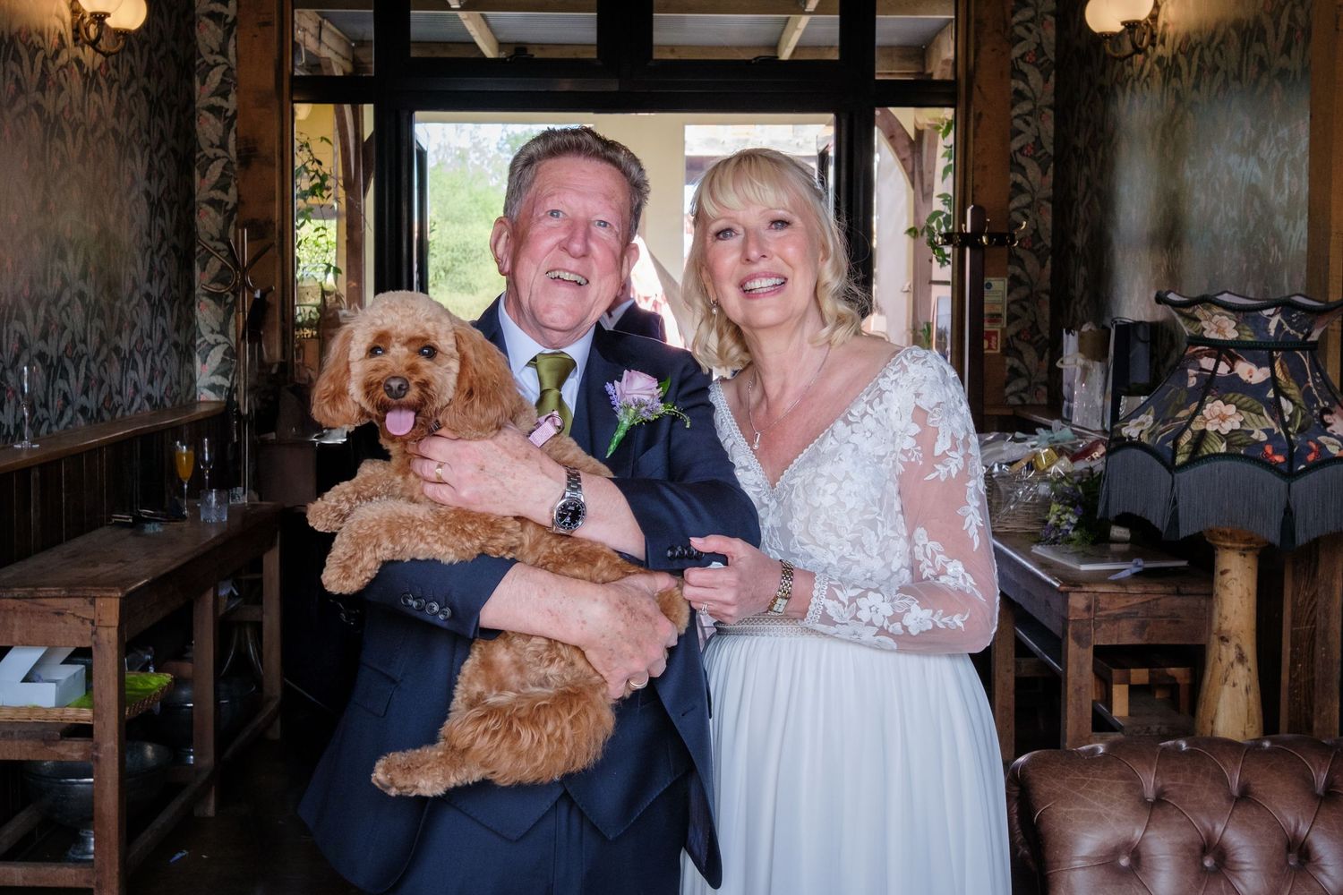 A wedding couple poses with their two small brown dogs at a rustic venue with wooden furniture and vintage decor.
