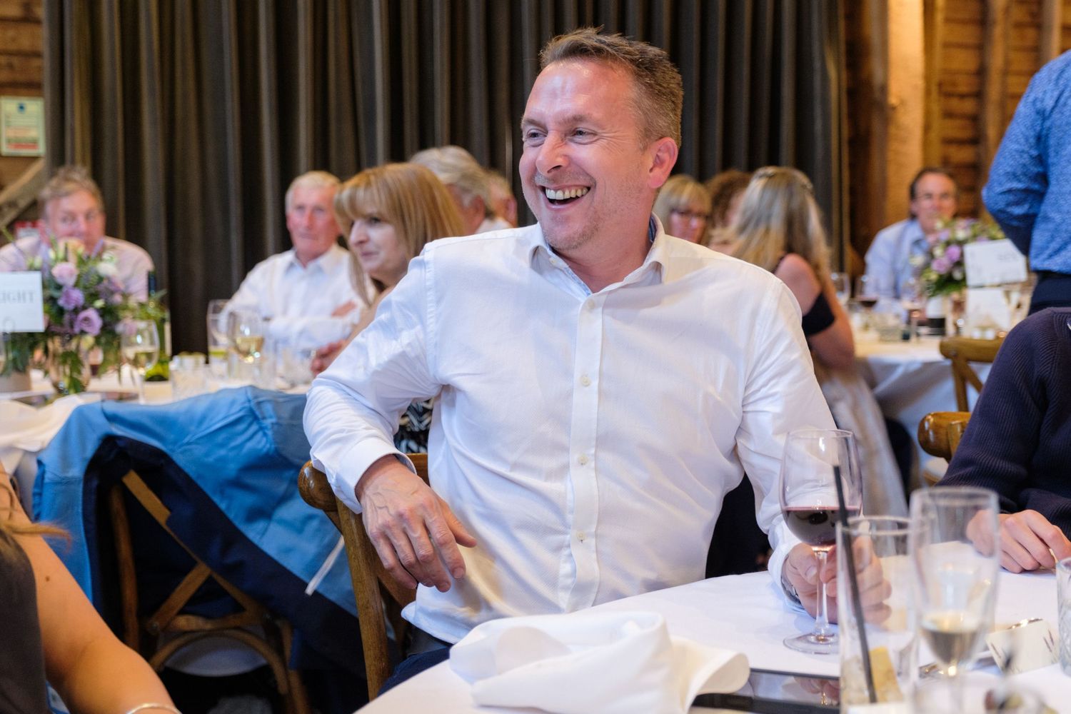 A person in a crisp white shirt laughs heartily during a dinner gathering at a restaurant with other guests visible in background.