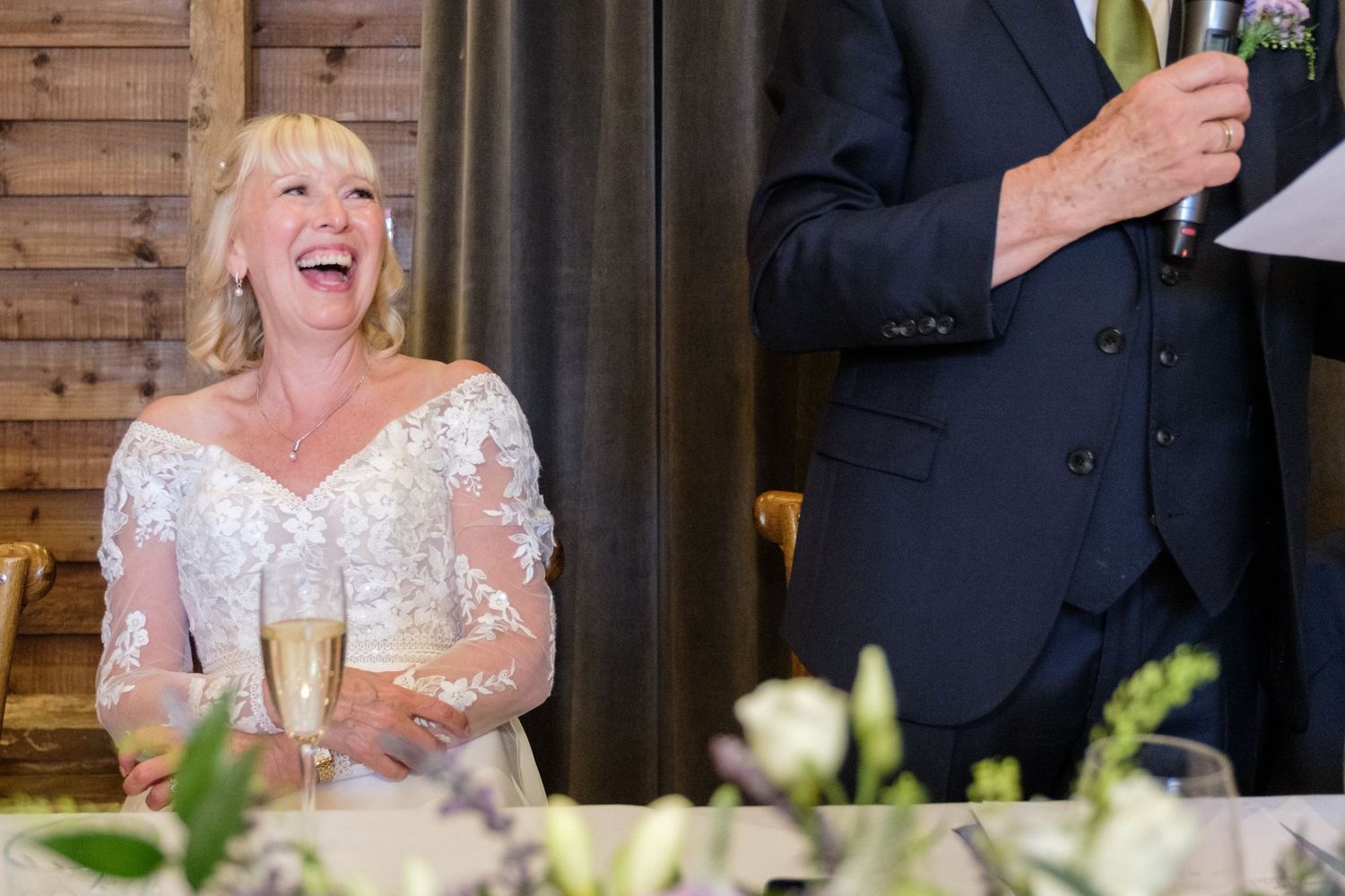 Joyous bride laughs during wedding reception toast while seated at table with champagne glass and floral decor.