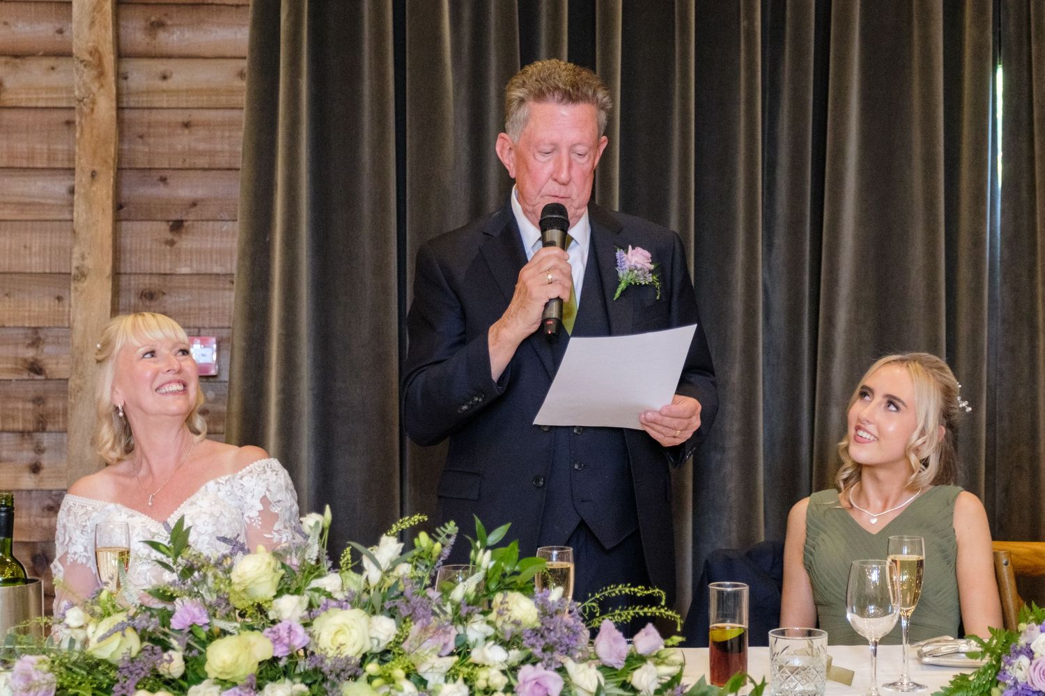 A speaker delivers a wedding speech at the head table while others listen alongside elegant floral arrangements and table settings.