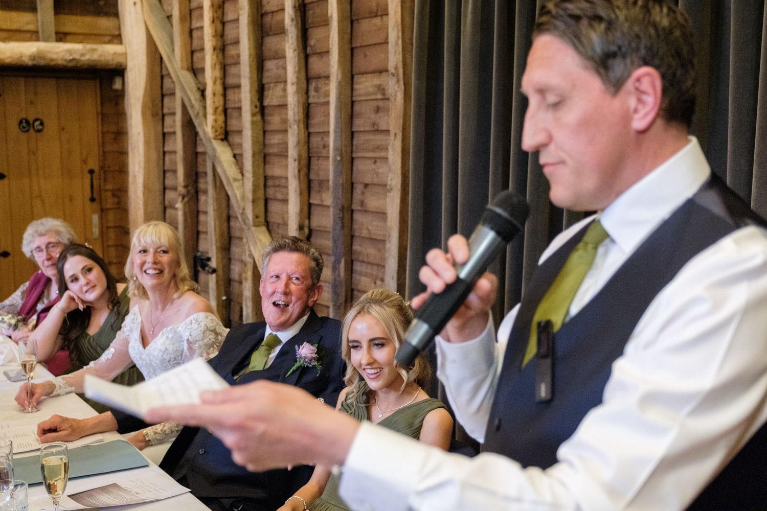 A wedding speaker in a white vest and green tie reads from papers at a head table during a reception.