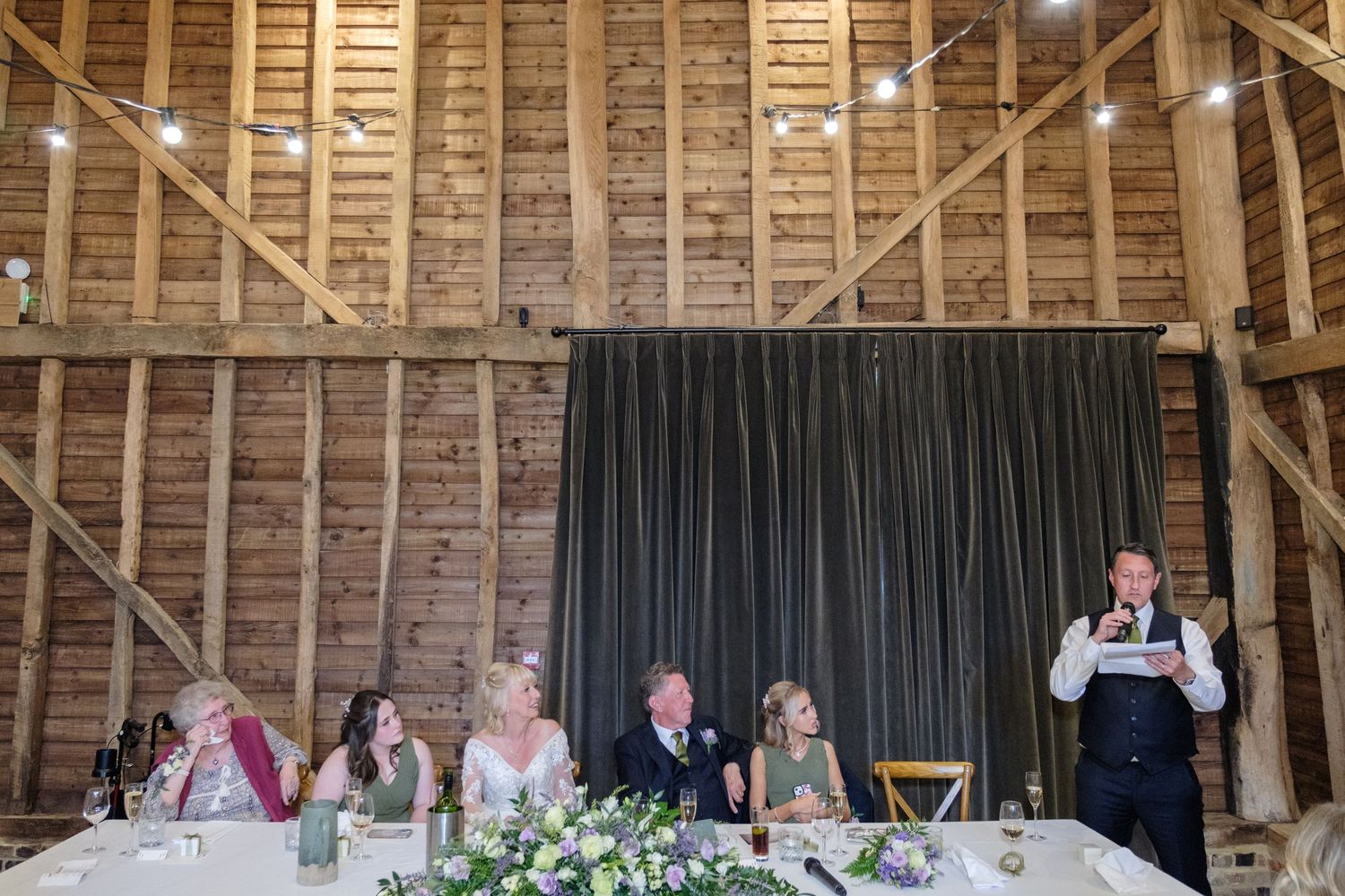 A wedding reception scene inside a rustic wooden barn with people seated at a head table and black curtain backdrop.