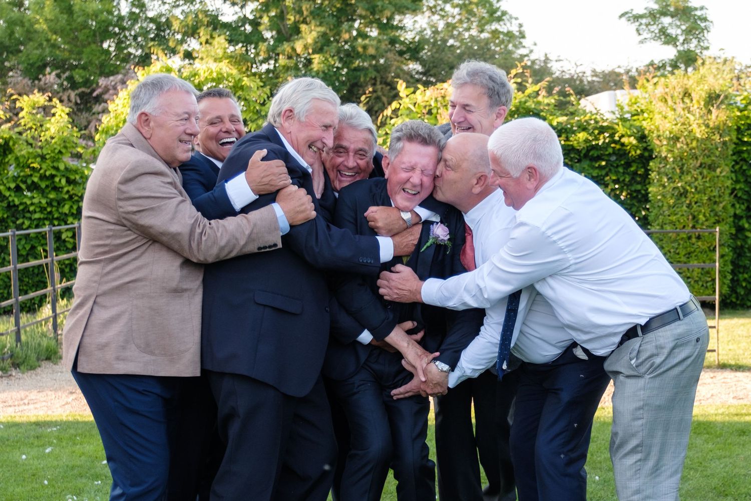 A group of older men in formal attire playfully huddle together outdoors in a garden setting.