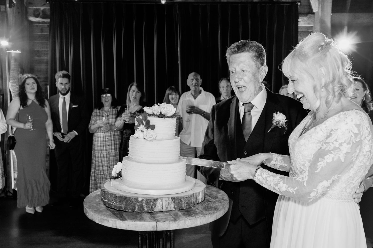 A black and white photograph of newlyweds cutting their wedding cake together at their reception while guests look on.