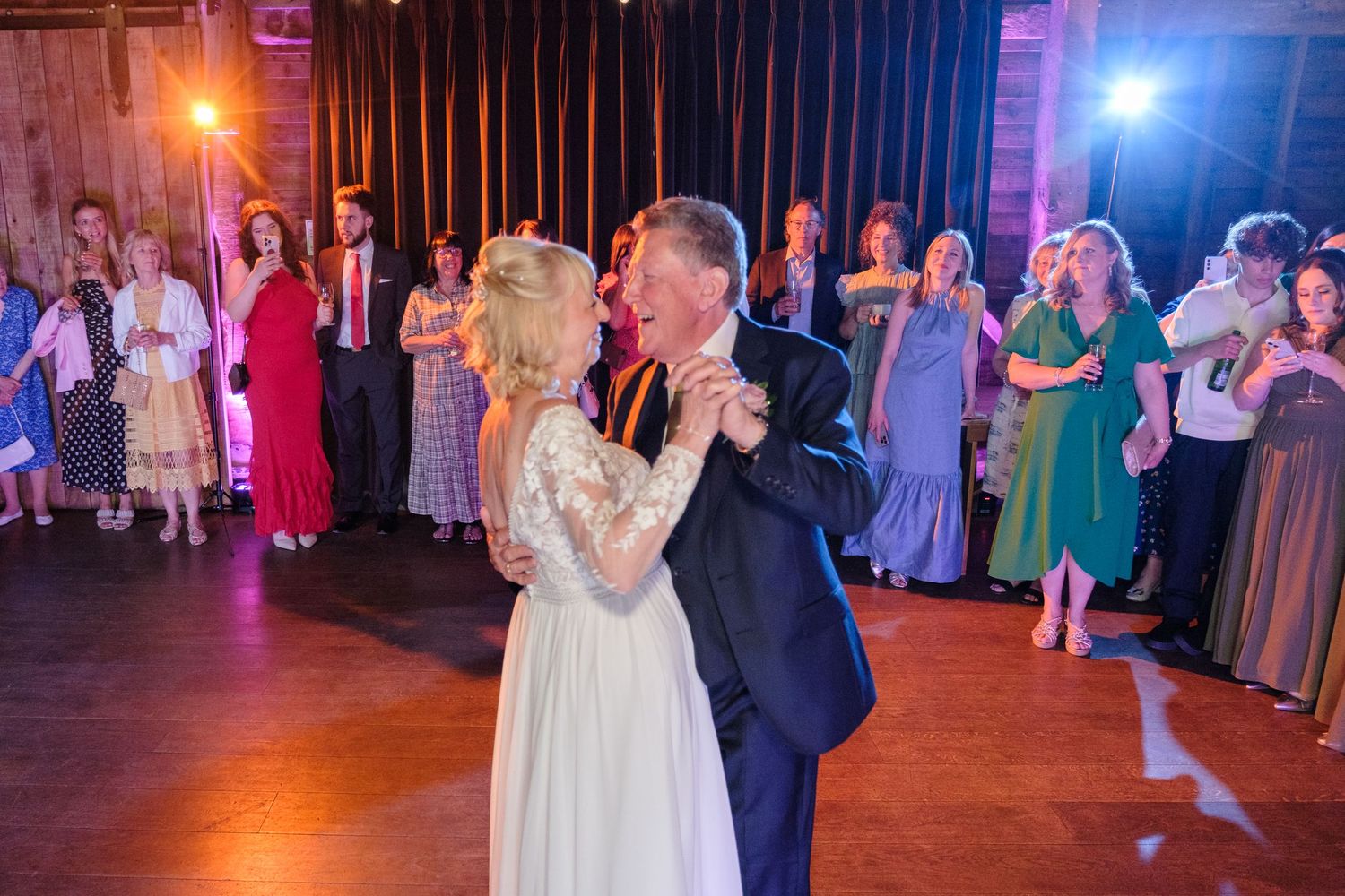 A couple dances together at a reception while guests watch from the sides under warm lighting on a wooden dance floor.