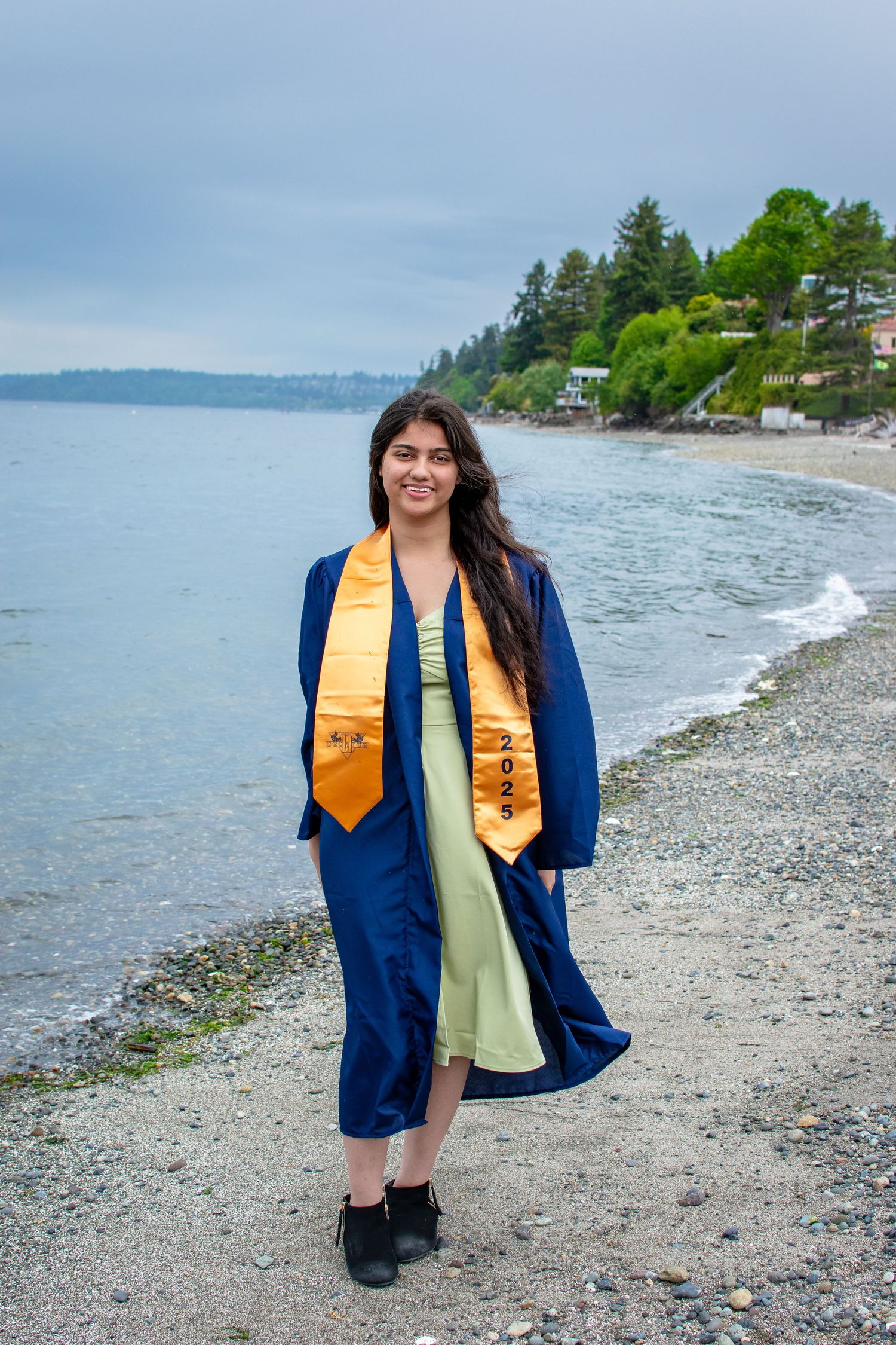 Class of 2025 graduate walking along the shoreline at Redondo Beach in Federal Way, WA in cap and gown.