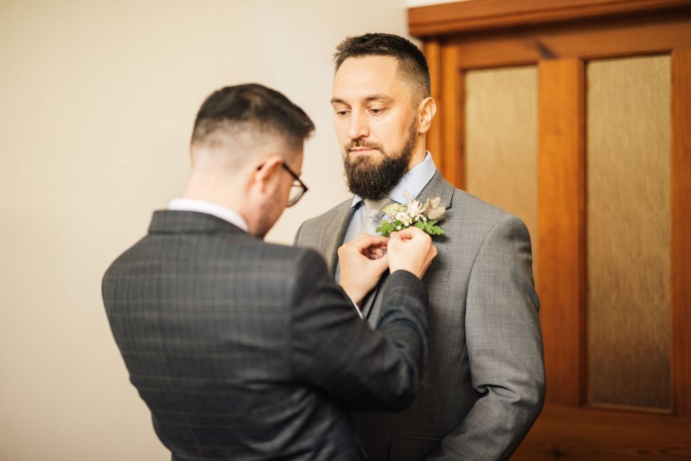 Boutonniere being pinned on grey suit jacket during wedding preparations.