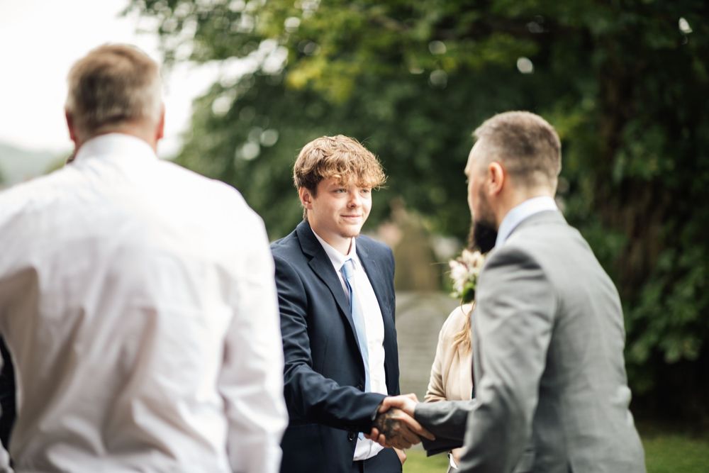 Two men in suits shake hands during an outdoor gathering while another person observes the interaction.
