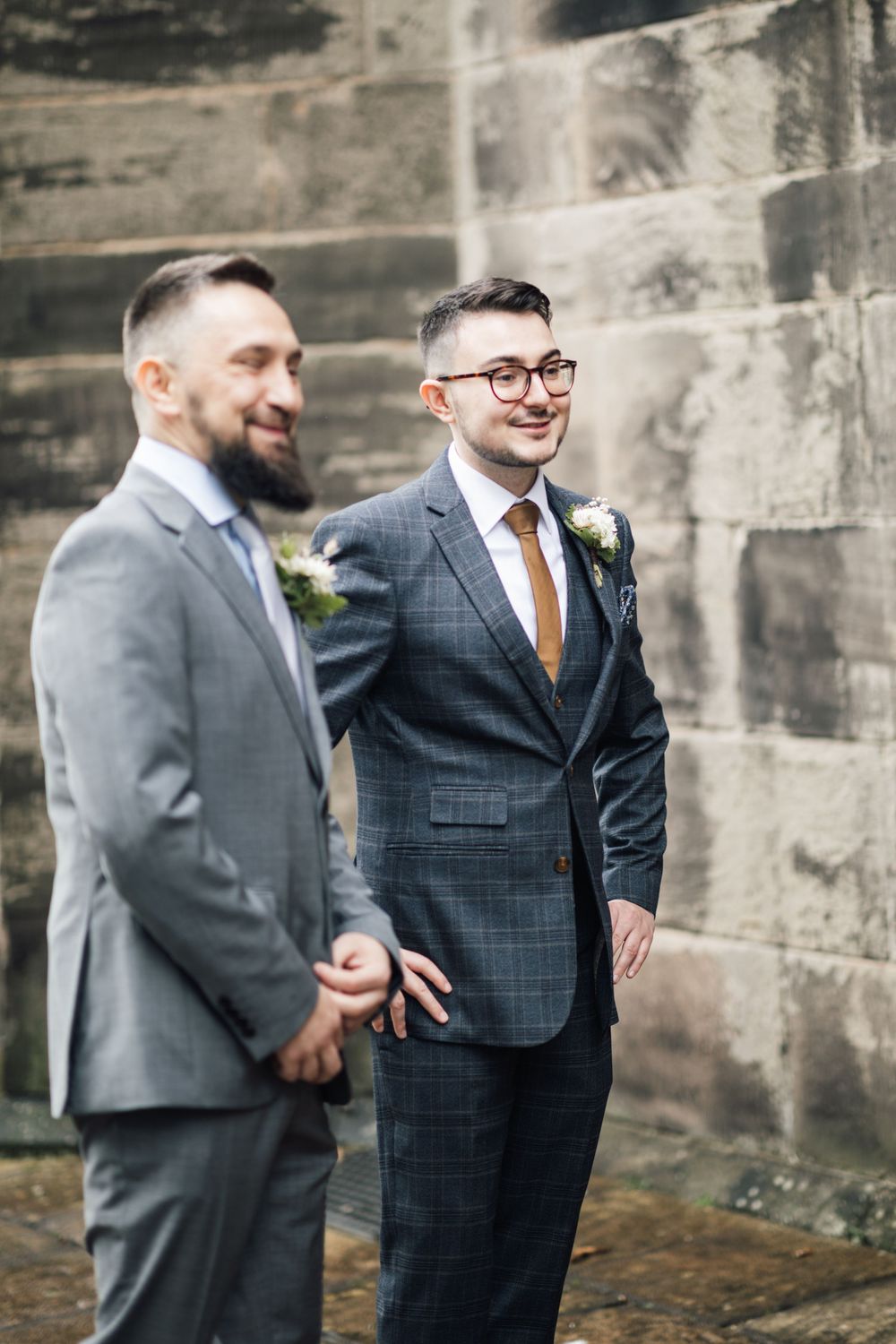 Two grooms in stylish suits share a joyful moment against a stone wall background during their wedding.