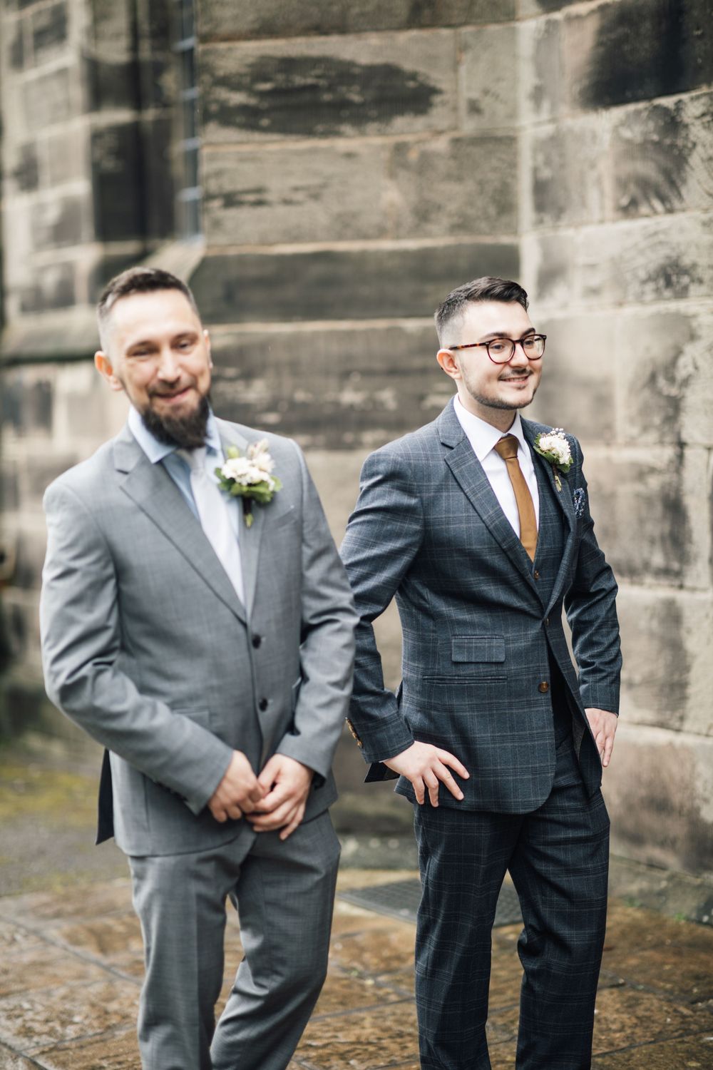 Wedding party members in contrasting gray and navy suits with boutonnieres stand against stone wall.