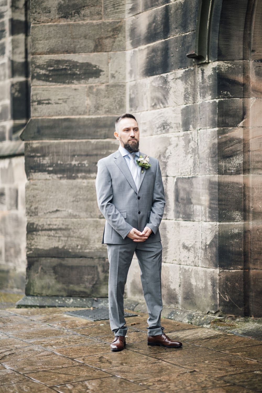 A person in a tailored gray suit stands outside a stone church building on a wedding day.