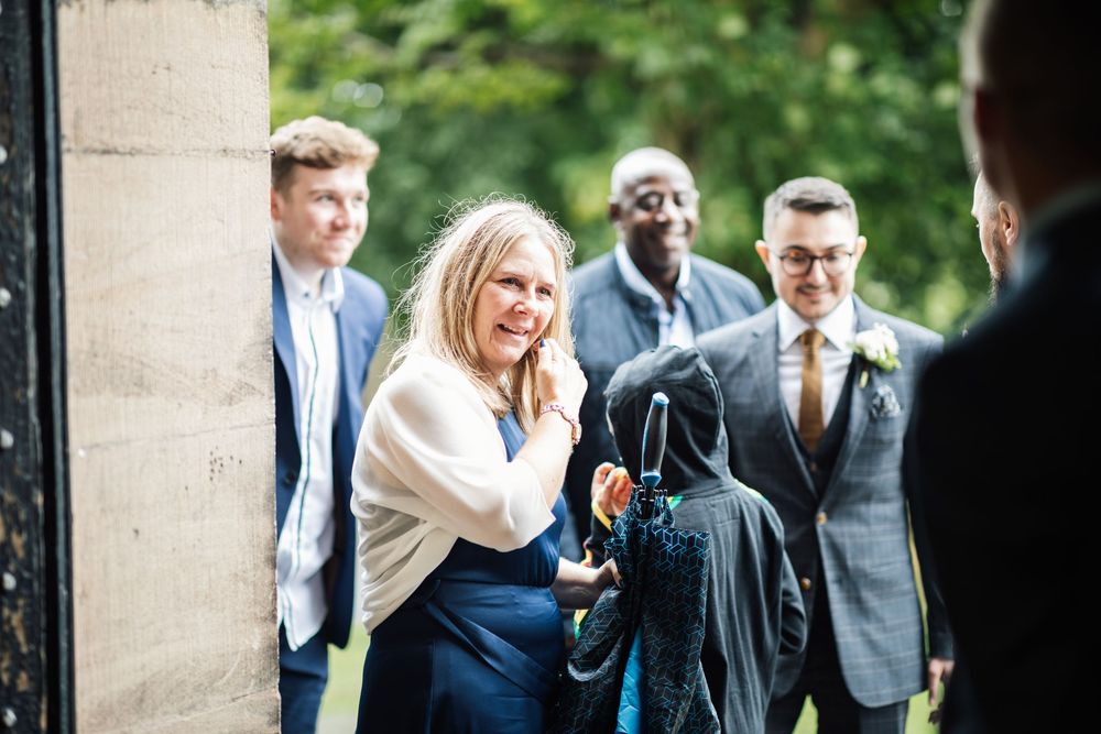 Wedding guests share a candid moment of laughter and joy outside a stone building on a sunny day.