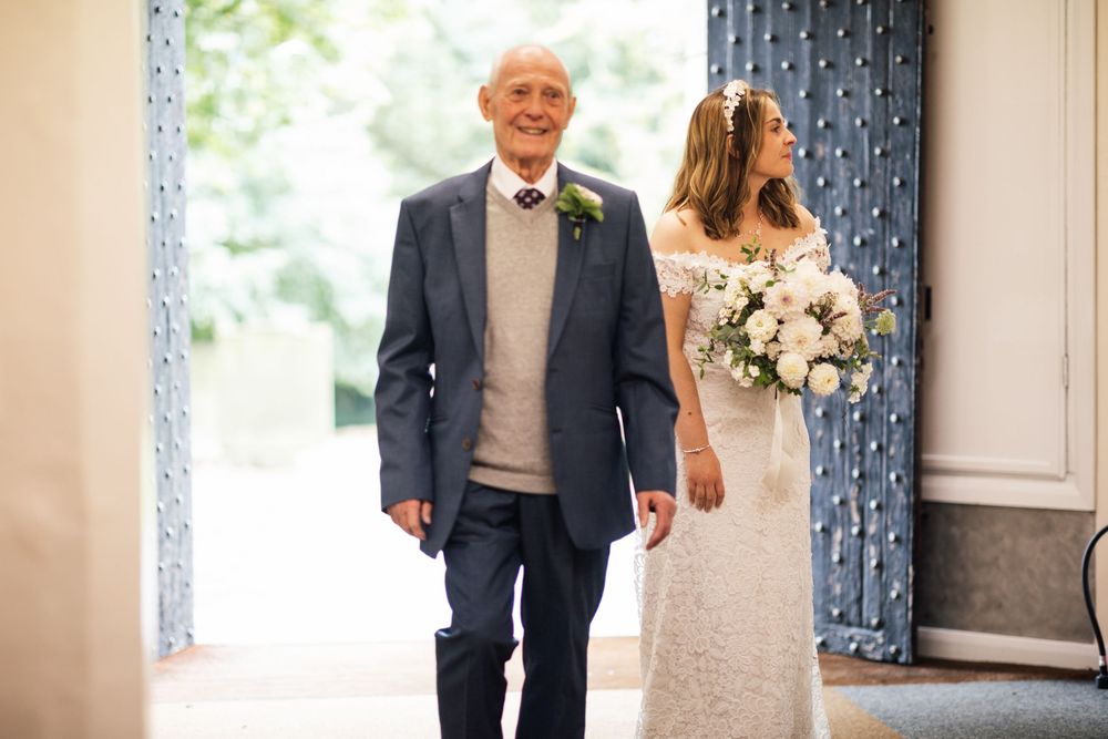 A person in formal attire walks down a bright hallway wearing a boutonniere while carrying a large white bouquet.