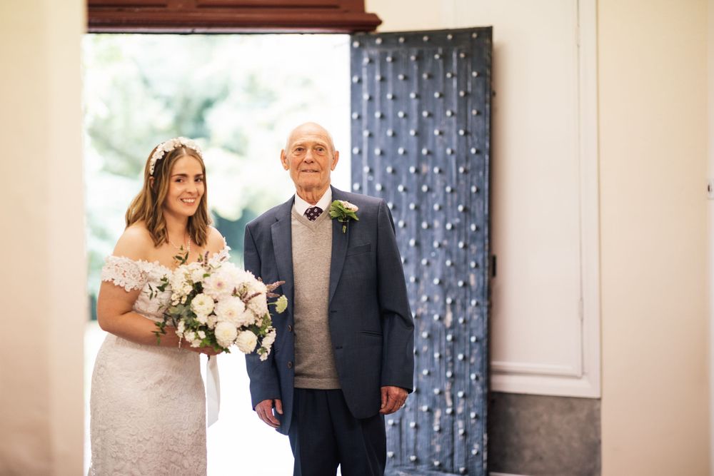 Two people stand together at a doorway before a wedding ceremony, one in a white dress holding a floral bouquet.