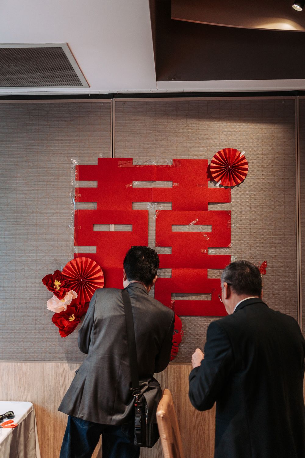 Two people in suits view traditional red Chinese wedding decorations with paper fans on a gray wall at an event.