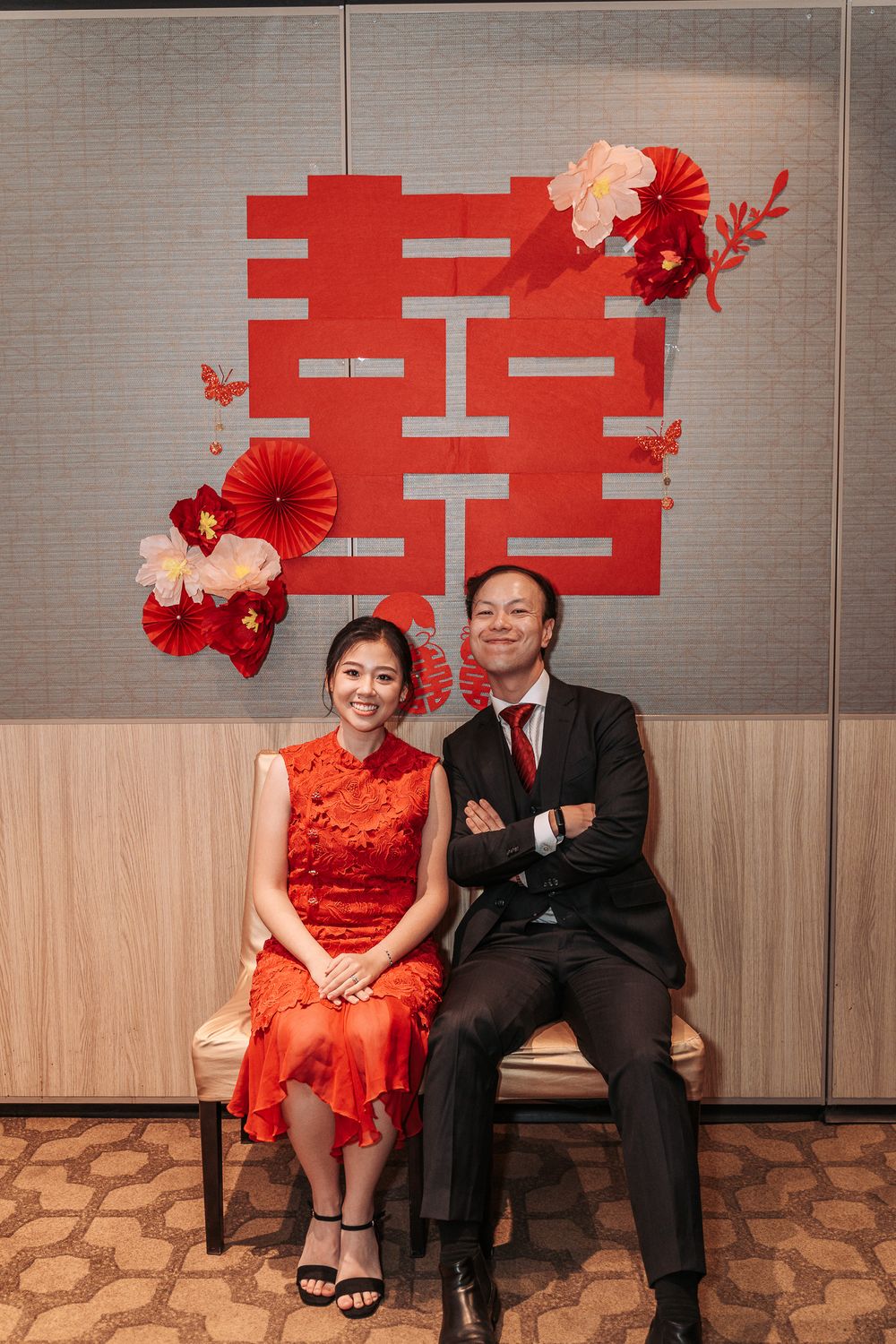 Traditional Chinese wedding scene with couple in formal attire seated against red double happiness symbol and floral decor.