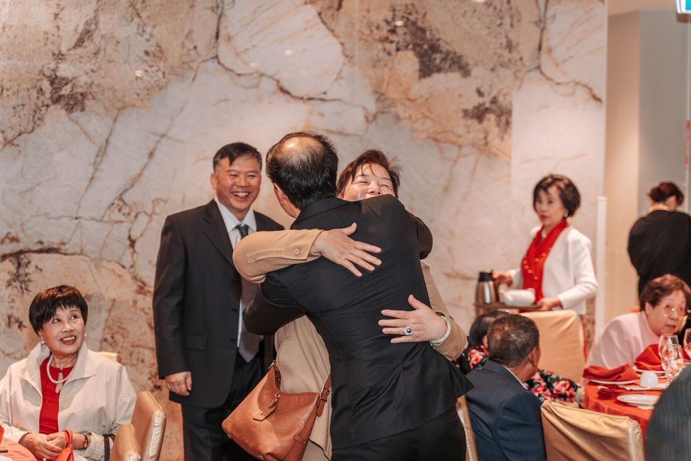 People embrace during a celebration or gathering in a room with marble-textured walls as others look on.