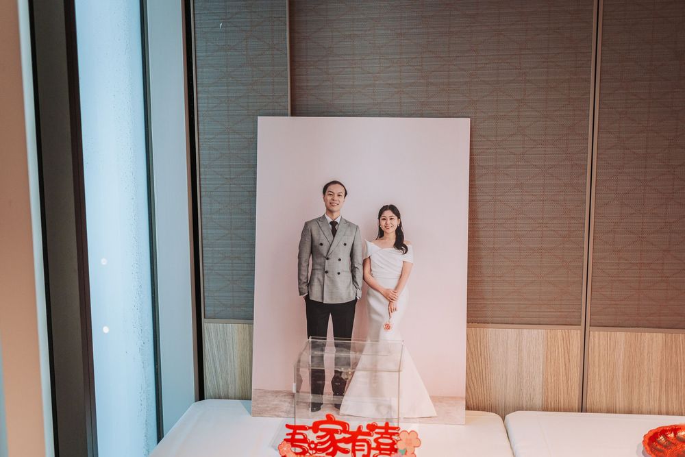 Two people stand in front of a white backdrop in a modern room with red petals scattered on the floor.
