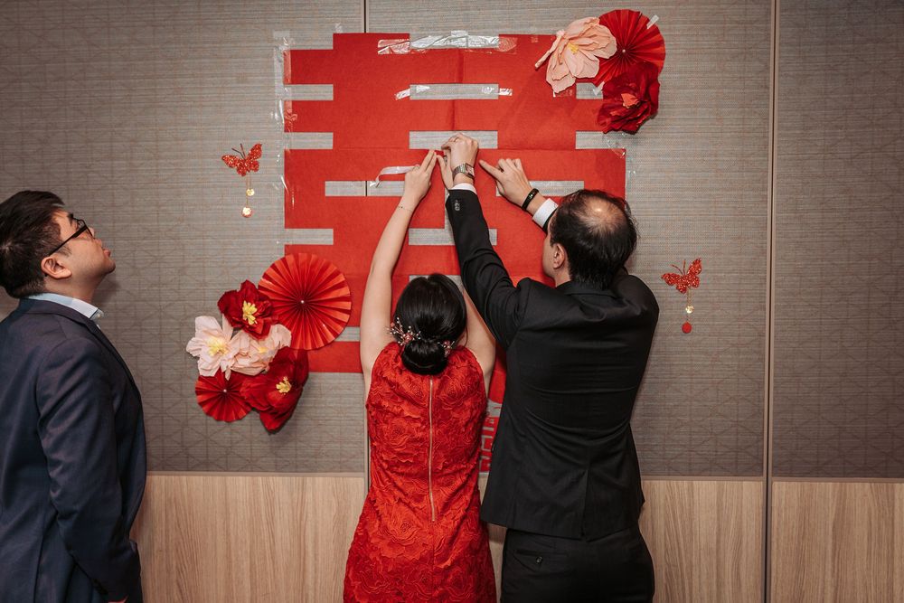 People in formal attire decorate a wall with red Chinese characters and traditional paper fans for a celebration.