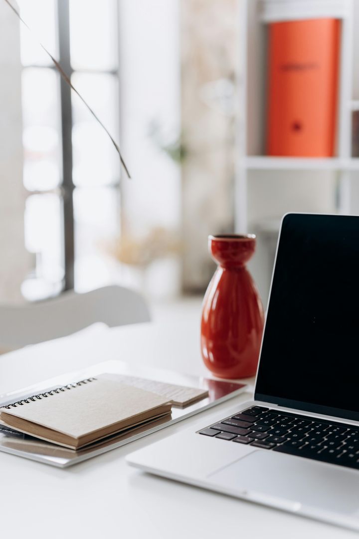 Bureau lumineux avec un ordinateur portable, un carnet et un vase rouge.