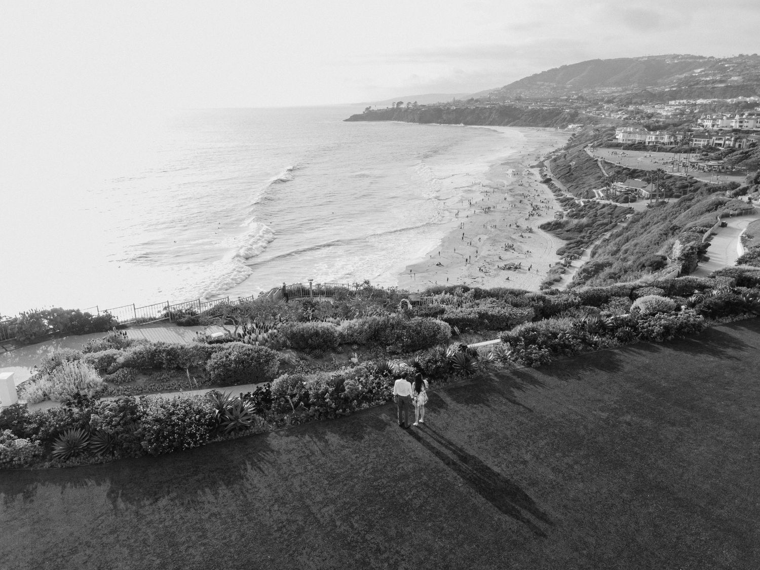 Black and white aerial view of a coastal landscape with rolling hills and curved shoreline along the ocean.