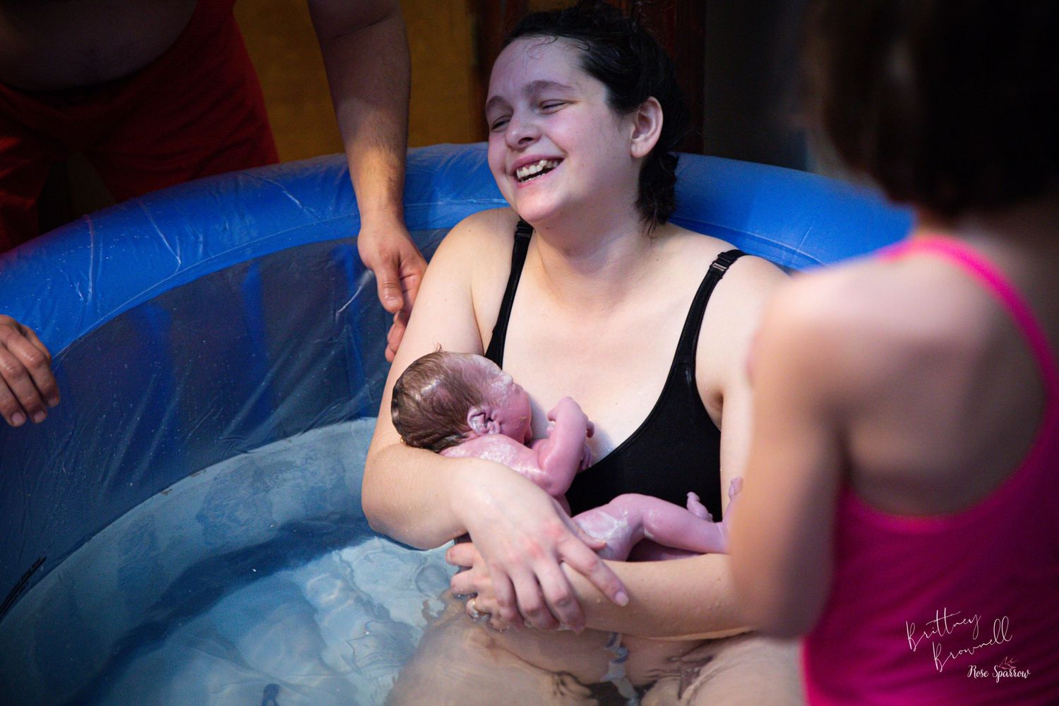 A water birth scene showing a newborn being cradled in a blue birthing pool while an older sibling takes in the scene.