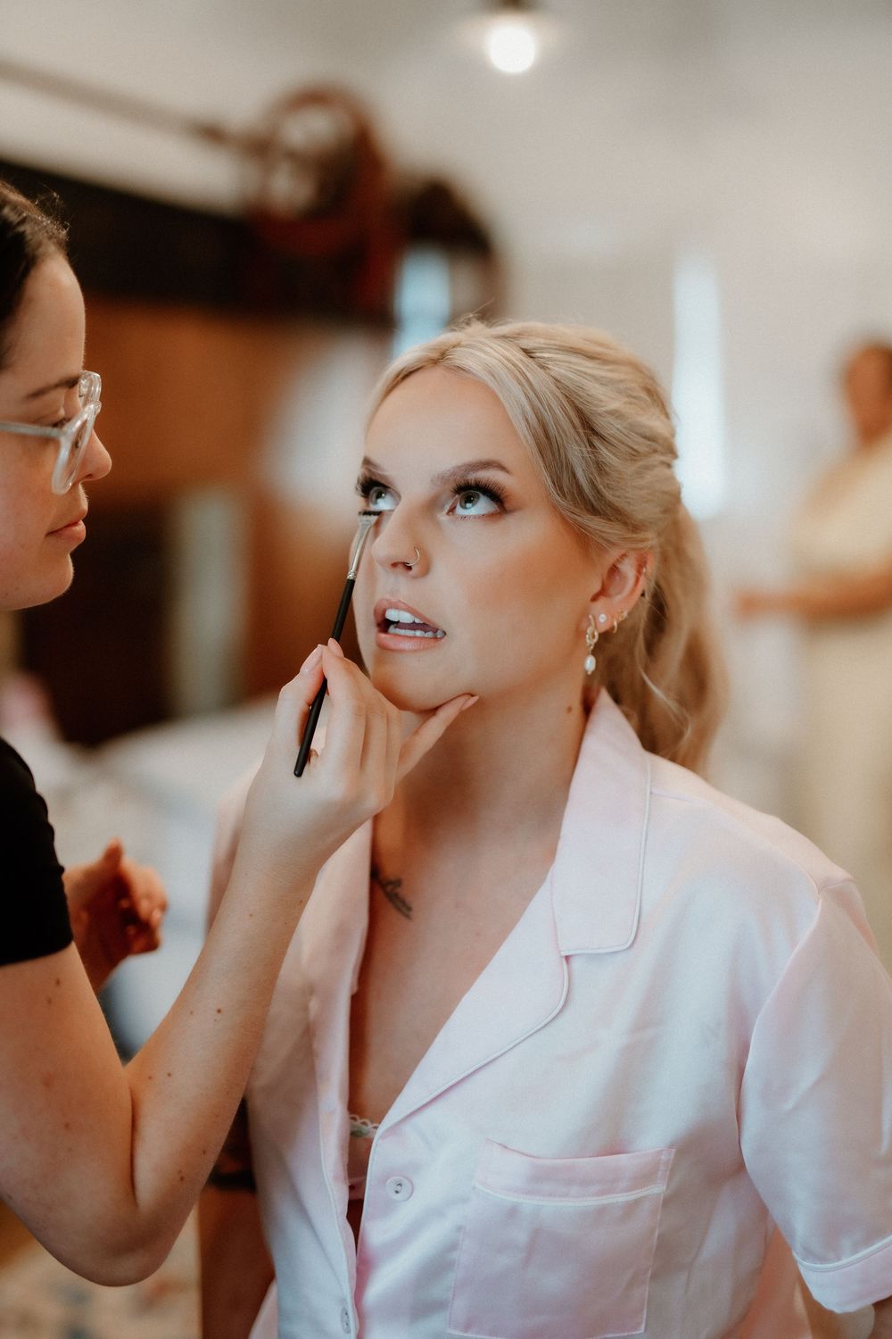 Makeup artist applies eye makeup on woman wearing white robe during wedding preparation.