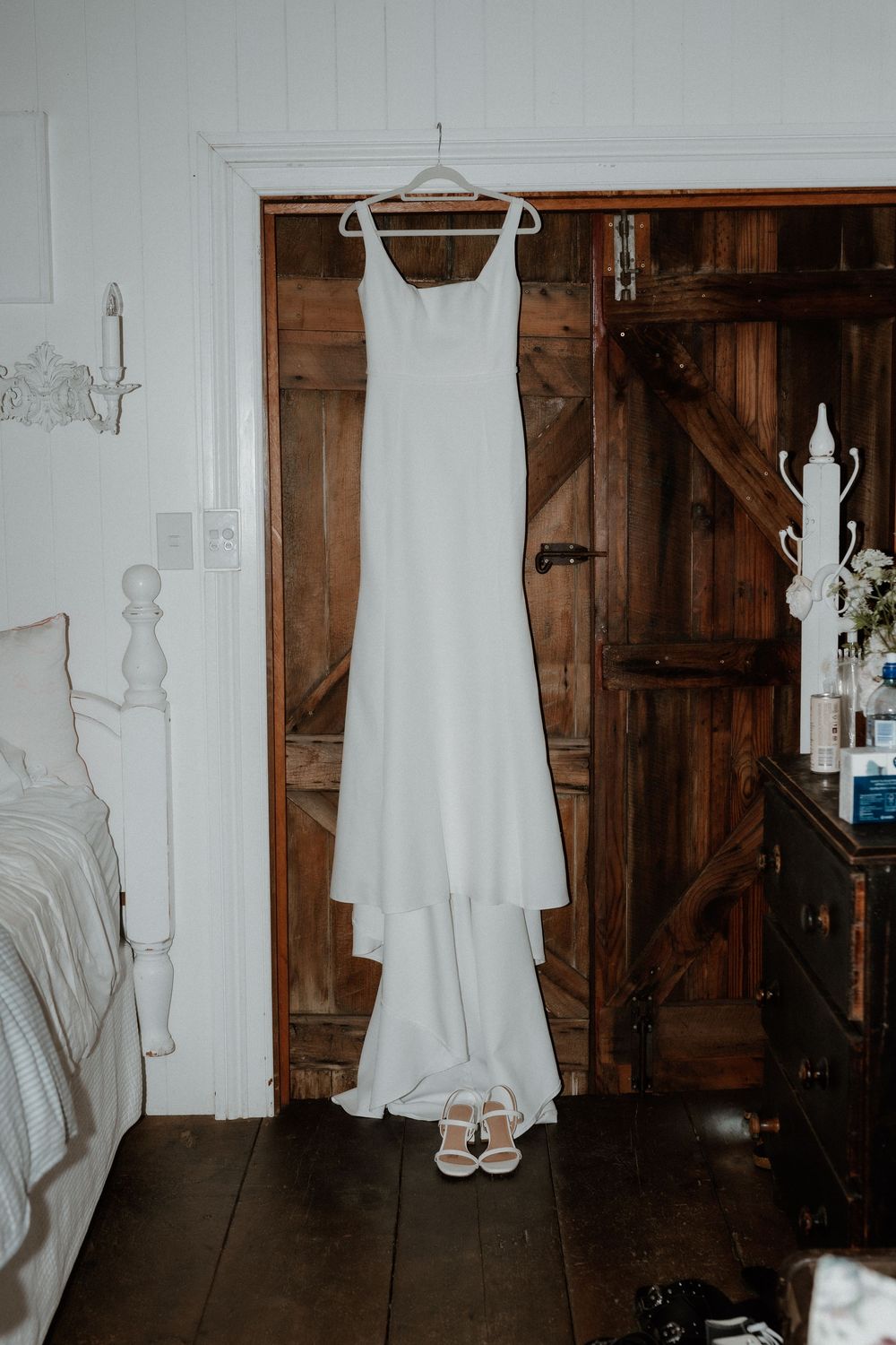Elegant white wedding dress and heels hanging on wooden door in rustic bedroom setting.