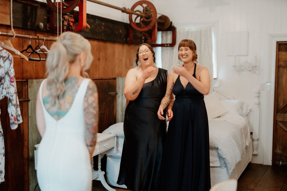 Bridesmaids in black dresses react excitedly to seeing bride in wedding dress.