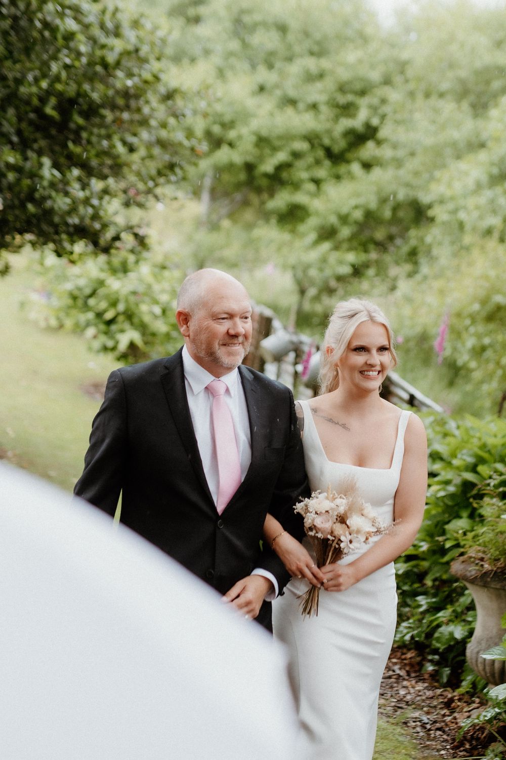 Two people walk arm in arm on outdoor garden path during wedding ceremony with lush greenery in background.