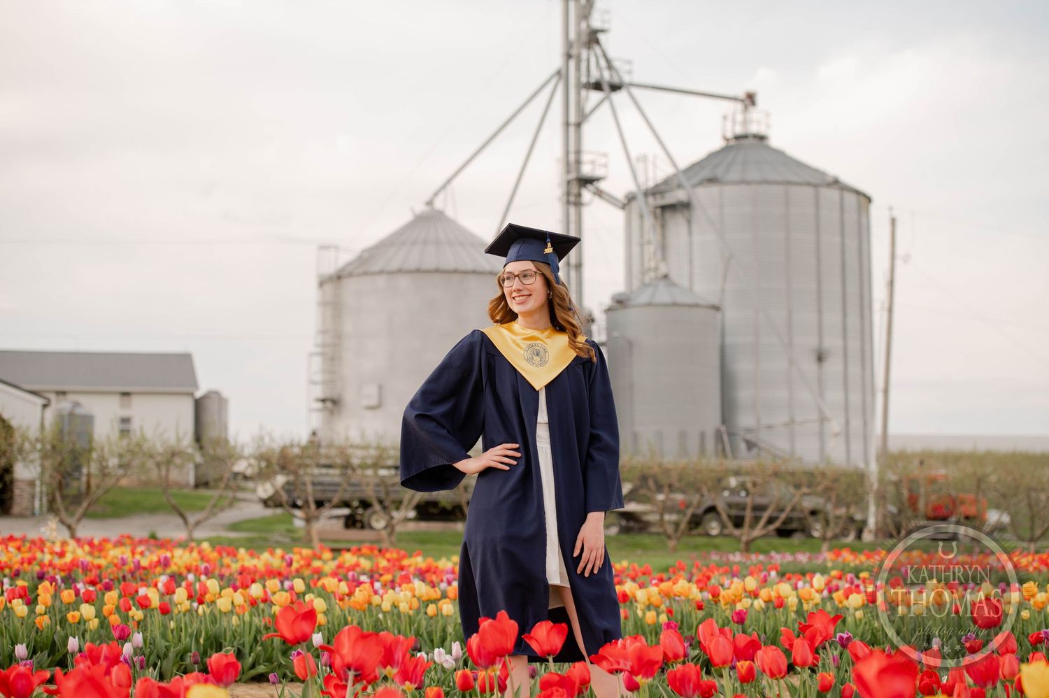 Graduation photo taken in a vibrant red tulip field with large grain silos in the background on an overcast day.