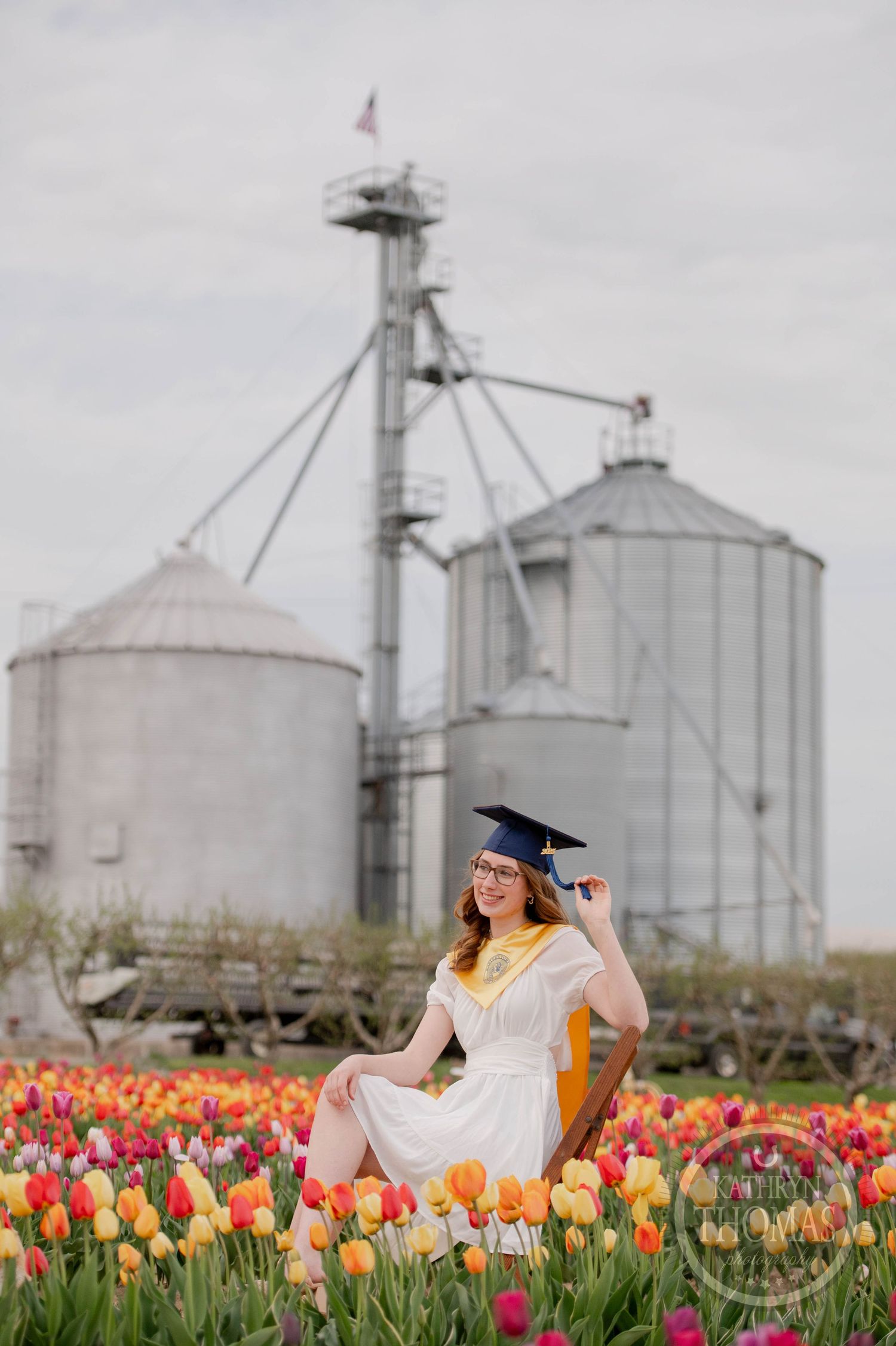Graduate in white dress sits among colorful tulips with grain silos and storage tanks in the background.
