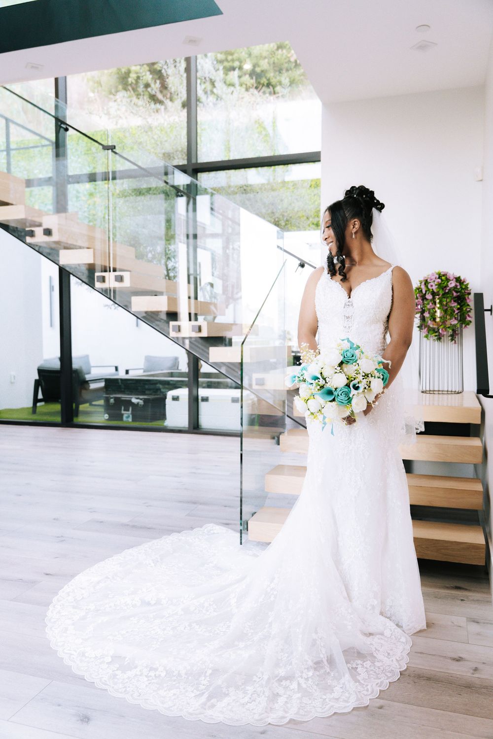 A bride in a flowing white lace wedding gown poses elegantly in a modern interior with glass staircase and wooden floors.