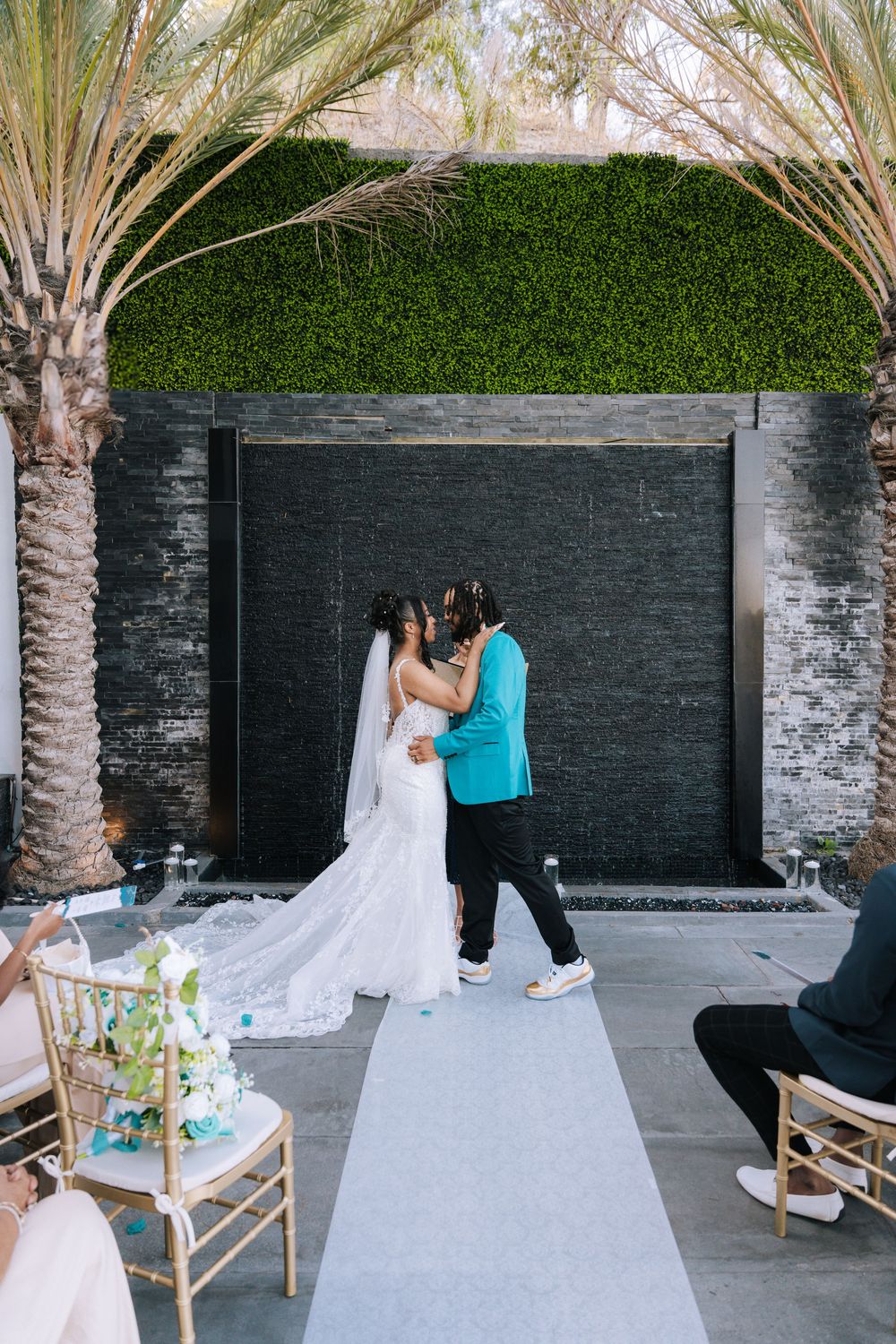 Wedding dress train being arranged and styled by assistants in an outdoor courtyard with palm trees and stone walls.