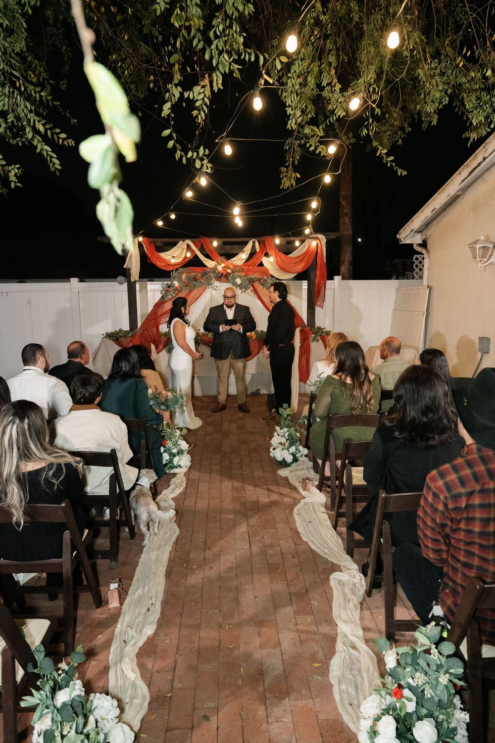 Intimate outdoor evening wedding ceremony with string lights, red draping, and floral aisle decorations on brick patio.