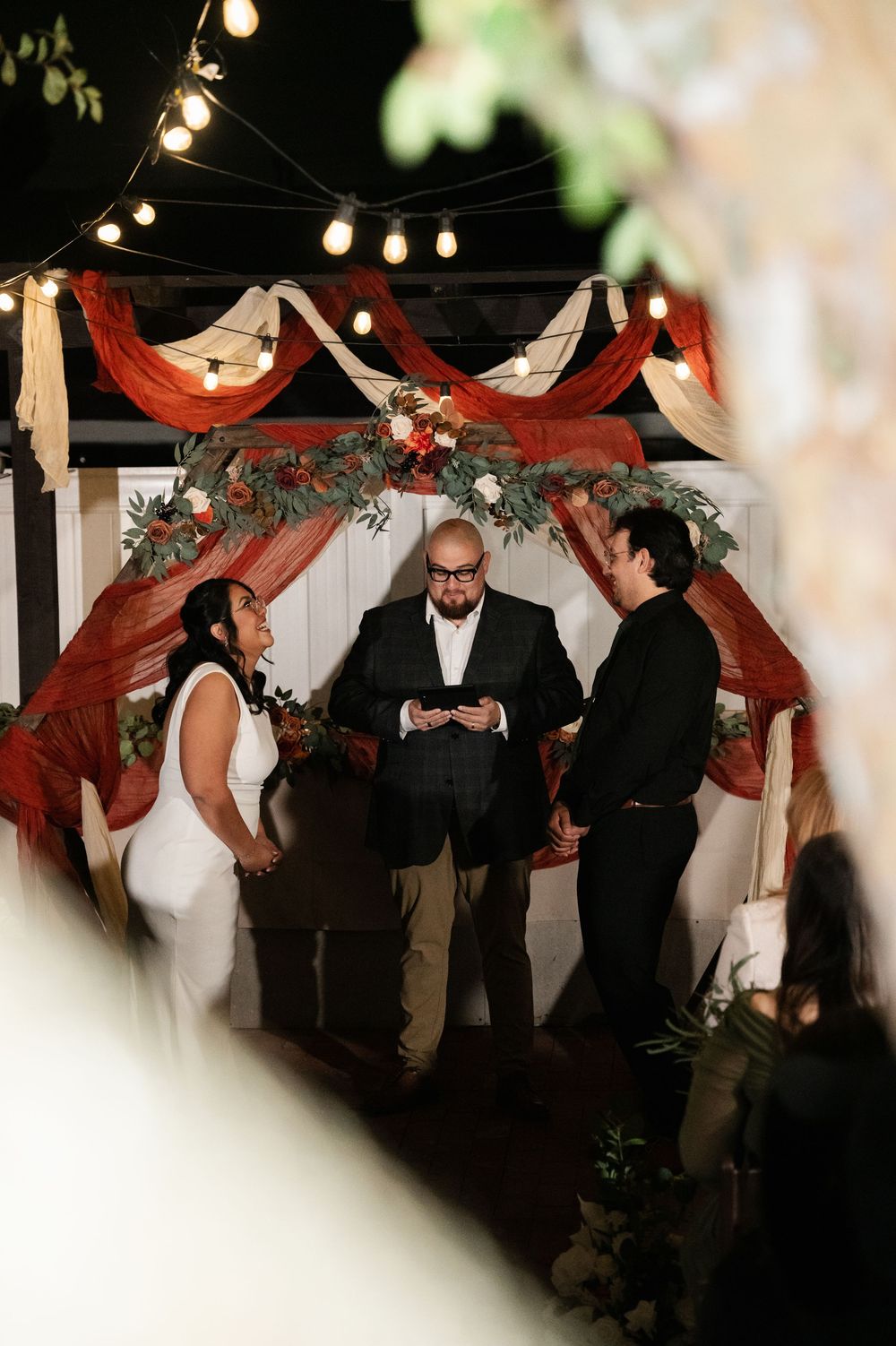 A wedding ceremony takes place under a draped red and white arch decorated with greenery and string lights overhead.