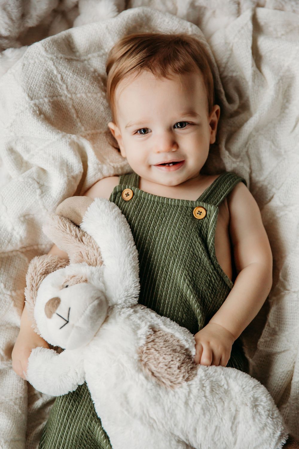 A smiling baby in green overalls cuddles with a plush lamb toy on cozy bedding.
