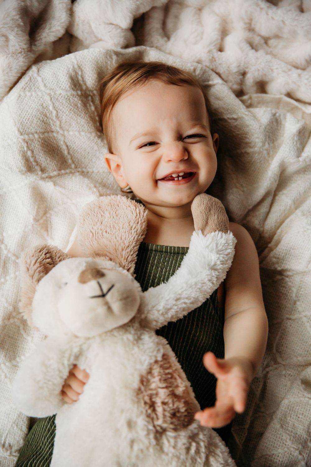 A joyful baby laughs while playing with a plush lamb toy on cream-colored blankets.