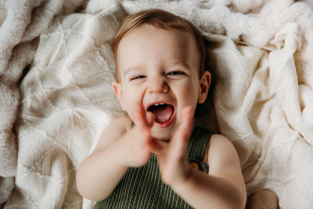 Baby wearing green knit romper lies on soft white blanket with joyful expression.