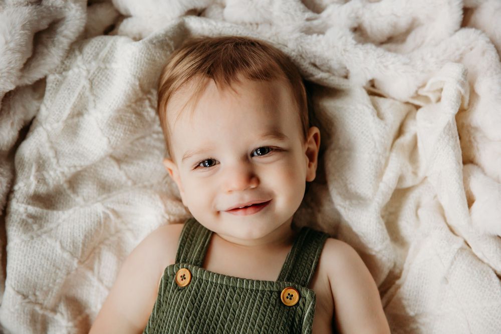 Smiling baby in olive green outfit rests on cream textured blanket.