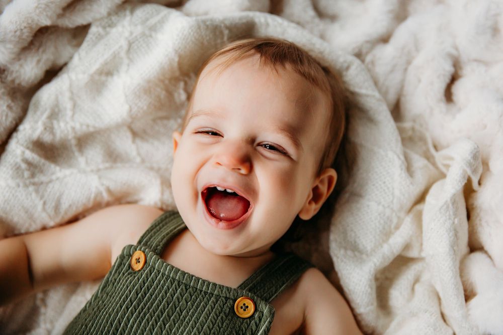 Laughing baby in dark green outfit plays on cozy white blanket.