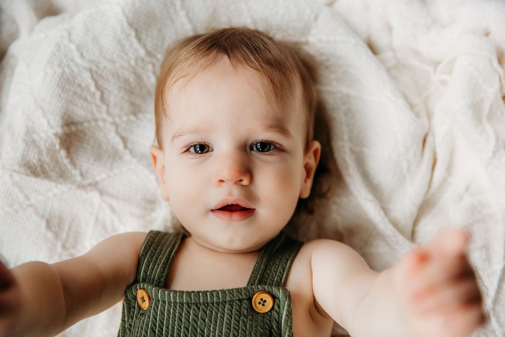 Infant wearing green romper with wooden buttons smiles on white textured surface.