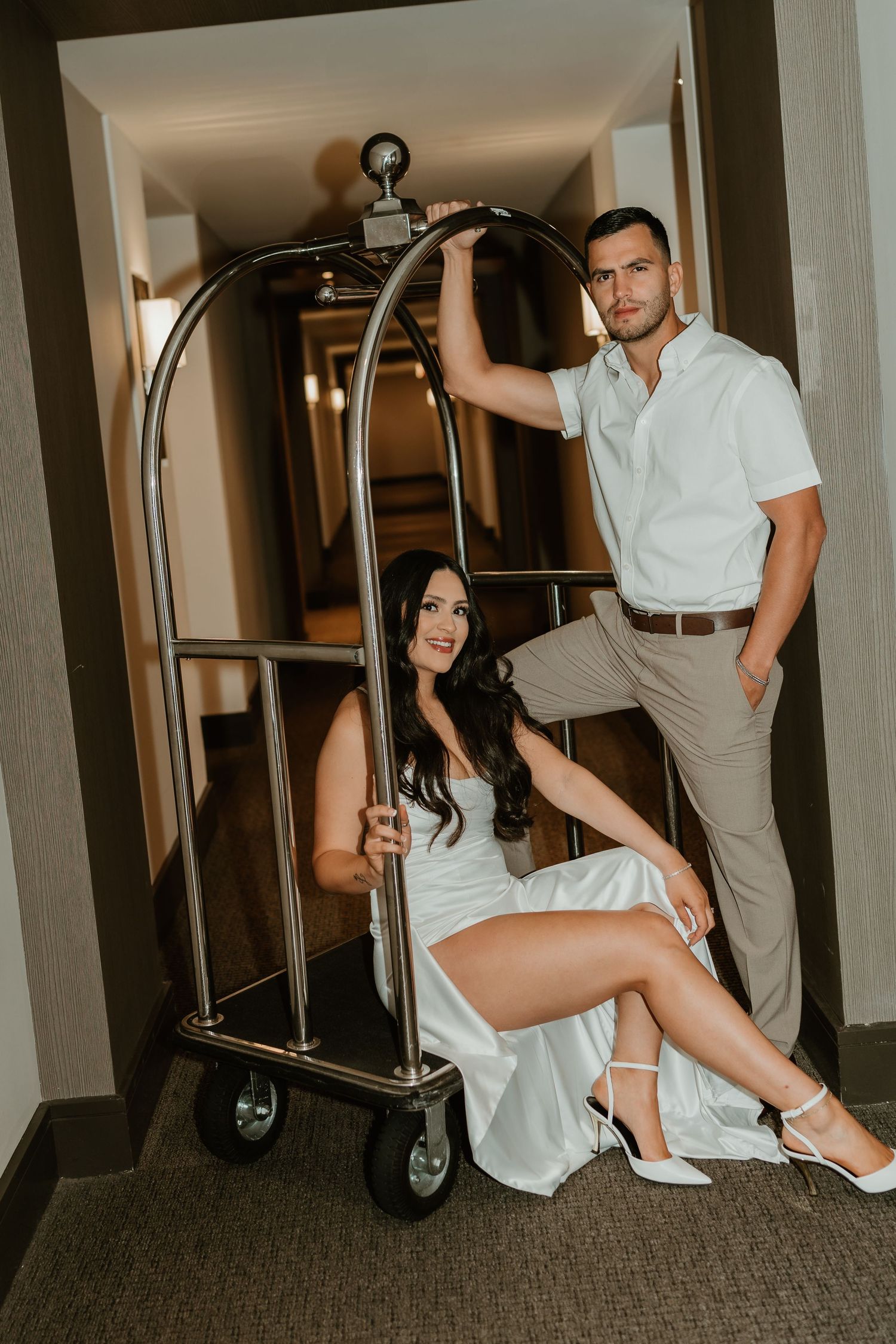 Couple in light colored outfits posing intimately in a hallway with dramatic lighting.