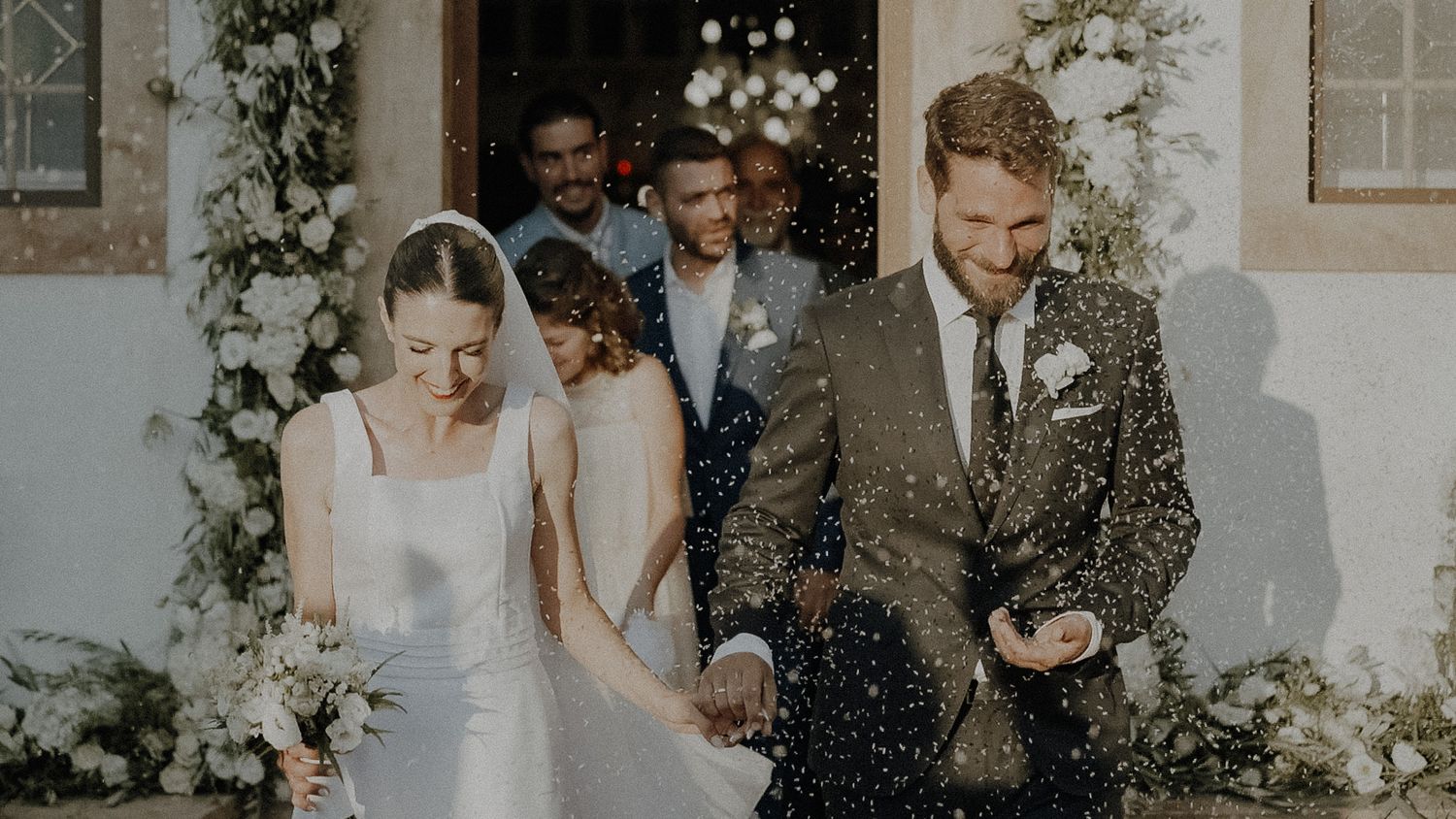 A joyful newlywed couple walks together through floating confetti outside a white building with floral decorations.