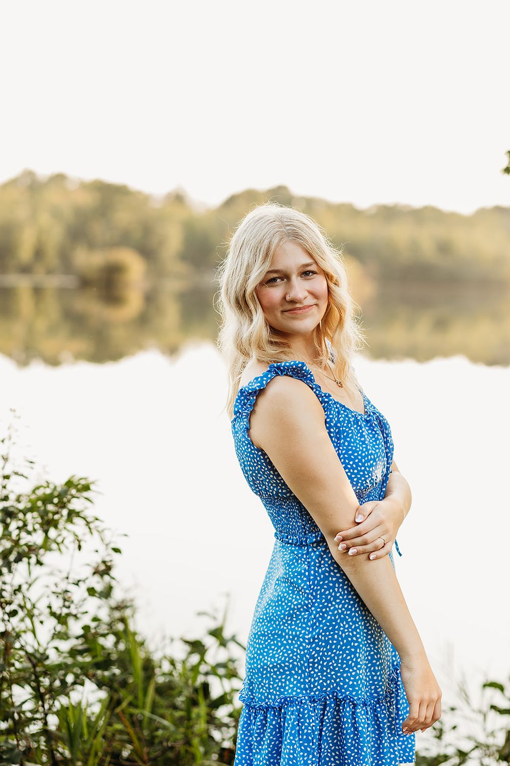 A woman in a blue polka dot dress smiles while standing by a tranquil lake at sunset.
